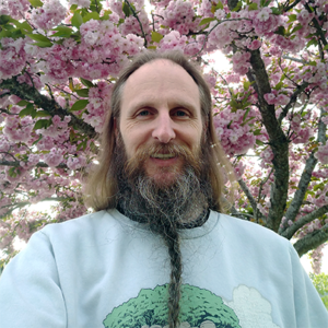 A white person with long hair, a long, braided beard and a moustache standing in front of a blooming cherry blossom tree.
